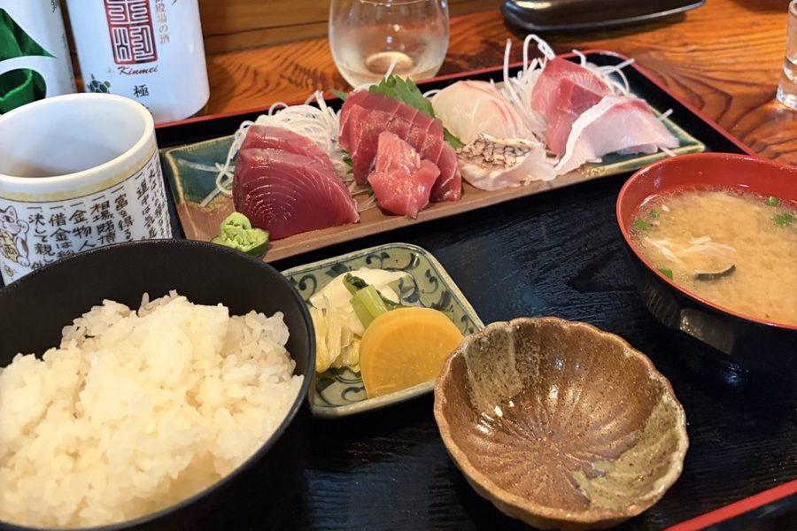 カウンターに置かれた刺身の盛り合わせの定食。ご飯、味噌汁、漬物、奥に地酒の瓶