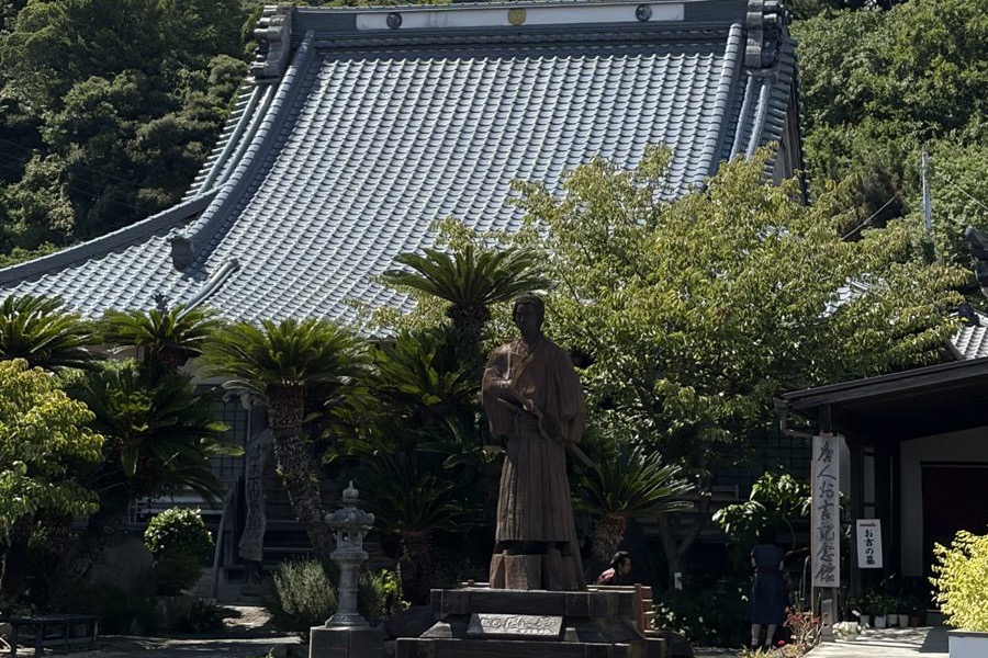 大きな瓦屋根の本堂の前に立つ銅像。まわりにソテツと石灯籠が並んでいる